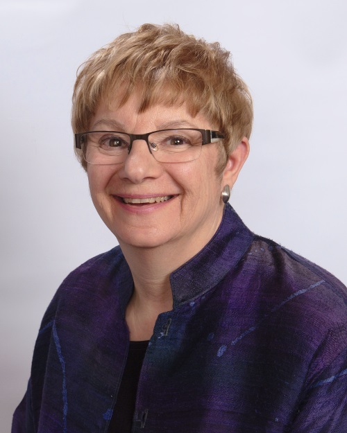 woman smiling. she has short hair, glasses and is wearing a purple jacket