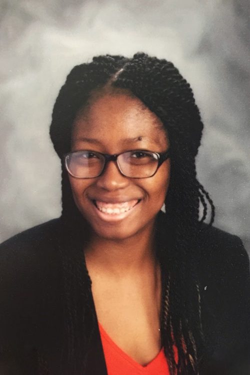 Woman: smiling, she is wearing glasses, a black blazer and a red shirt.