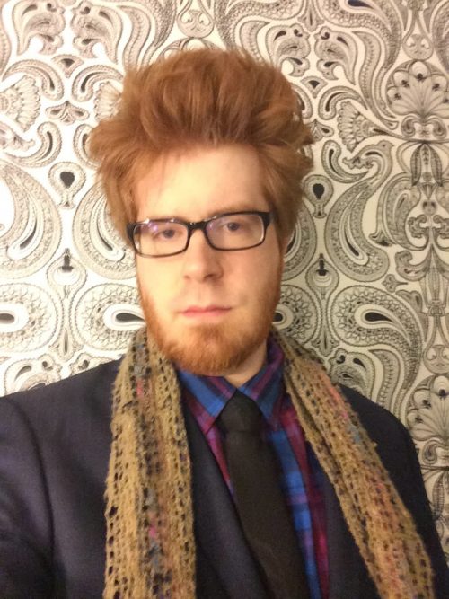 man with red hair, glasses and a beard wearing a dark suit and scarf