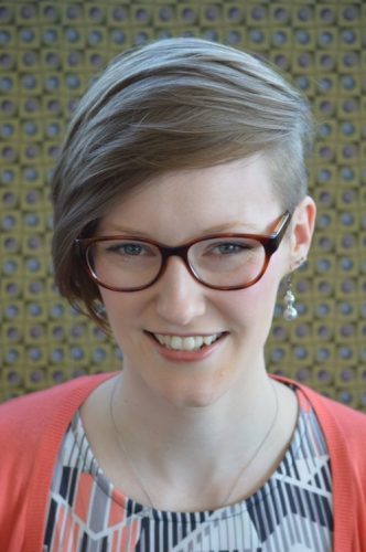 woman with light brown hair, glasses, wearing a patterned shirt and pink sweater.