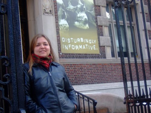 woman with shoulder-length brown hair, wearing a black leather jacket and red scarf, in front of a sign that says disturbingly informative